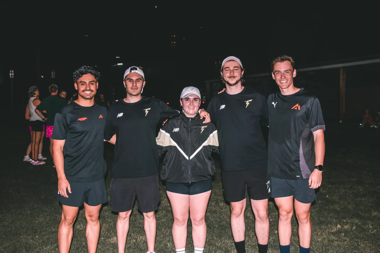 Five smiling runners in black club tops standing together at a night run, with a dark field behind them